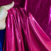 Une main tient un morceau de Lamé métallique liquide au mètre de Longan Craft France, avec une finition magenta brillante. D'autres pièces de ce tissu lamé extensible vintage pour les costumes et la couture sont suspendues à l'arrière-plan.