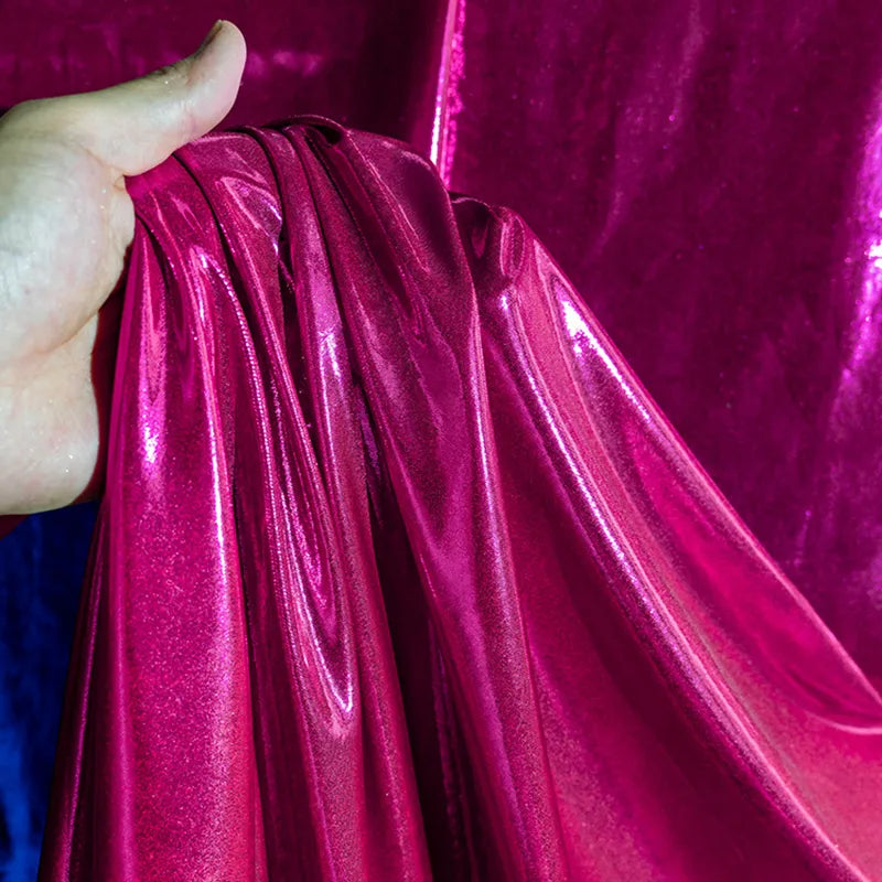 Une main tient un morceau de Lamé métallique liquide au mètre de Longan Craft France, avec une finition magenta brillante. D'autres pièces de ce tissu lamé extensible vintage pour les costumes et la couture sont suspendues à l'arrière-plan.