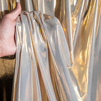 Une main présente une section de Lamé métallique liquide au mètre de Longan Craft France, un tissu extensible vintage brillant et réfléchissant pour les costumes, les robes et la couture, avec d'autres pièces de tissu suspendues à l'arrière-plan.