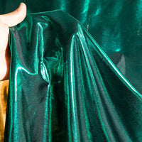 Une main présente le Lamé métallique liquide au mètre de Longan Craft France - un tissu lamé vintage brillant et extensible, d'un vert irisé, qui reflète la lumière pour mettre en valeur son drapé et sa texture remarquables - idéal pour les robes de soirée ou les costumes de scène.