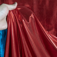Une main présente le lamé métallique liquide au mètre de Longan Craft France - un tissu vintage brillant et extensible pour les costumes, les robes et la couture - avec sa surface rouge réfléchissante et une touche de bleu en arrière-plan.