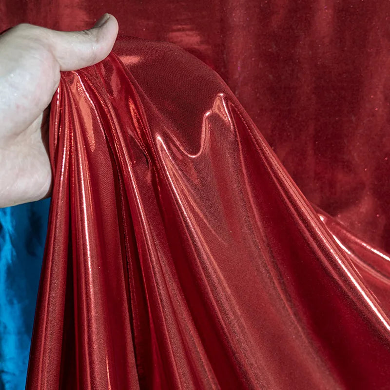 Une main présente le lamé métallique liquide au mètre de Longan Craft France - un tissu vintage brillant et extensible pour les costumes, les robes et la couture - avec sa surface rouge réfléchissante et une touche de bleu en arrière-plan.
