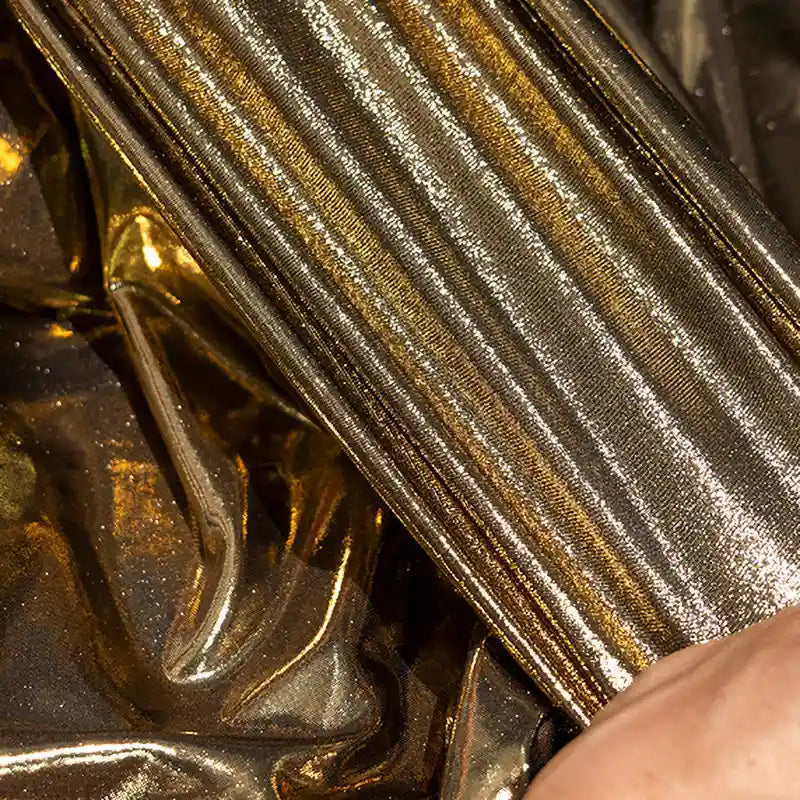 Une main touche le plissé, le brillant Lamé métallique liquide au mètre de Longan Craft France - un tissu vintage et extensible idéal pour les costumes et les robes de soirée - à côté d'un tissu métallique doré froissé.