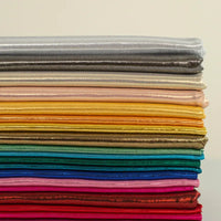 Une pile horizontale de Lamé métallique liquide au mètre de Longan Craft France brille en argent, or, jaune, vert, bleu, rose et rouge - parfait pour les costumes de scène, les robes et la couture - sur un fond uni.