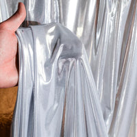 Une main soulève un morceau de Lamé métallique liquide au mètre de Longan Craft France devant une toile de fond argentée. Ce tissu lamé brillant et extensible est idéal pour les costumes de scène, les robes de soirée et la couture.