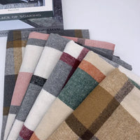 Six écharpes pliées en Tissu en flanelle à carreaux pour l'automne (polyester-coton) de Longon Craft France, dans les coloris marron, beige, gris, vert, rose et blanc, évoquent la douceur de l'automne lorsqu'elles sont présentées sur une surface blanche.