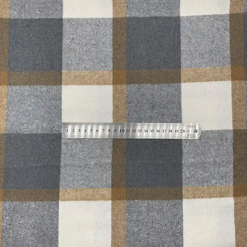 Une règle en plastique transparente de 20 cm repose sur le "Tissu en flanelle à carreaux pour l'automne au mètre" de Longon Craft France, un tissu écossais doux en polyester et coton, parfaitement aligné avec ses carreaux gris, bruns et blancs.