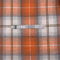 Une règle en métal, marquée en centimètres, est posée sur un tissu en flanelle à carreaux pour l'automne au mètre de Longon Craft France - un tissu à carreaux orange, gris et blanc en polyester-coton, idéal pour les peignoirs, les chemises et les projets de couture.