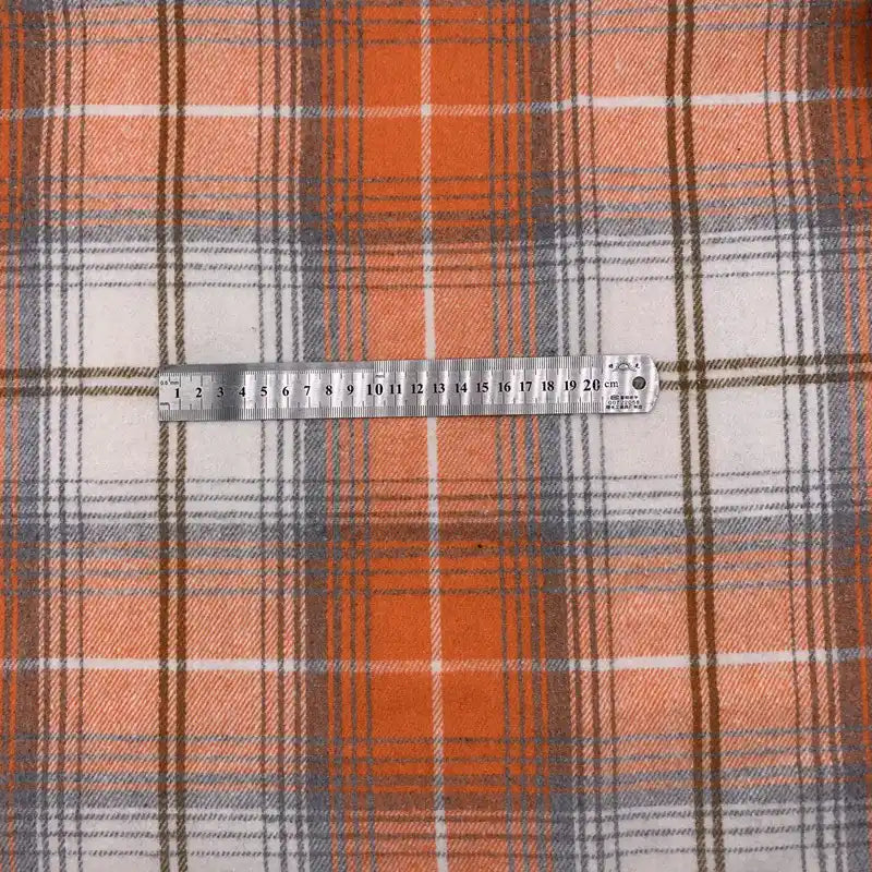 Une règle en métal, marquée en centimètres, est posée sur un tissu en flanelle à carreaux pour l'automne au mètre de Longon Craft France - un tissu à carreaux orange, gris et blanc en polyester-coton, idéal pour les peignoirs, les chemises et les projets de couture.