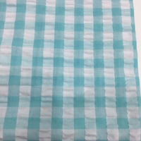 Un gros plan sur le tissu seersucker à carreaux au mètre en vichy turquoise et blanc de Longan Craft France met en évidence son quadrillage régulier et sa surface texturée - cette matière légère est idéale pour les vêtements d'été.