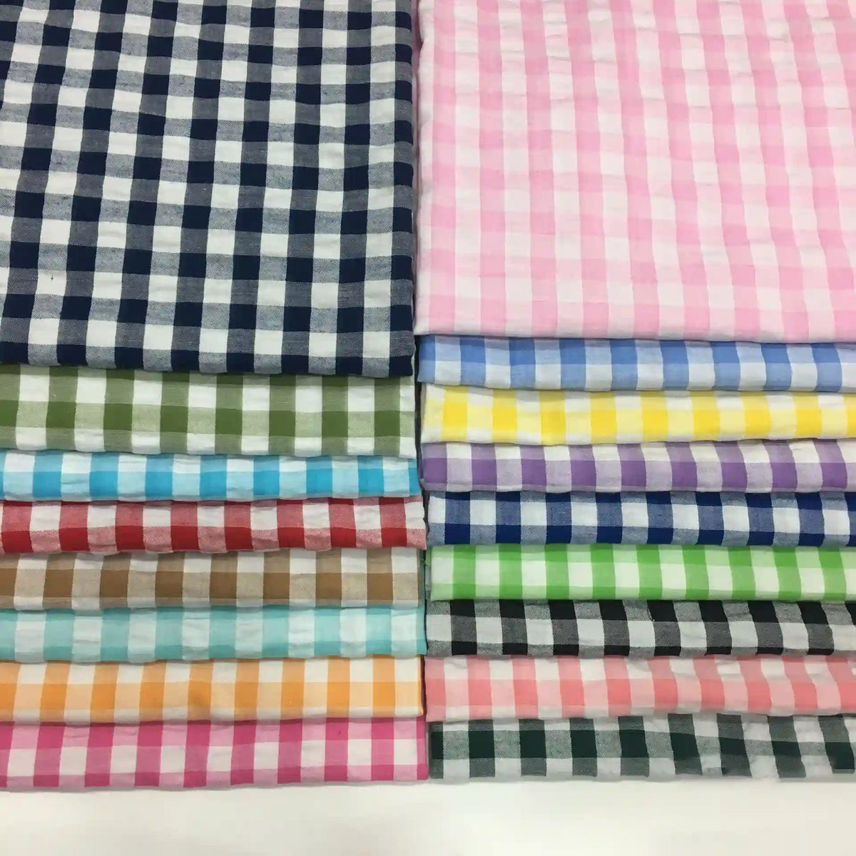 Des tissus seersucker à motif vichy de Longan Craft France, soigneusement empilés : colonnes à gauche en tons foncés, à droite en couleurs claires et pastel-idéals pour des vêtements d'été légers.
