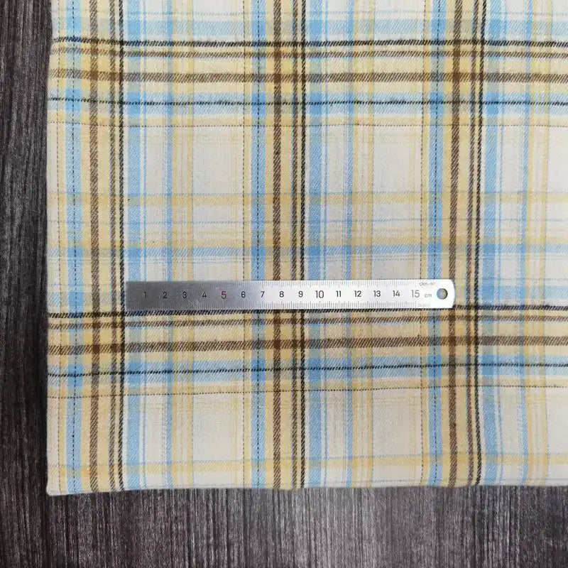 Une règle en métal repose sur le tissu flanelle à carreaux polyester-coton au mètre de Longon Craft France, un tissu d'automne douillet idéal pour les vêtements de bébé, avec un motif jaune, bleu et beige sur une surface en bois foncé.