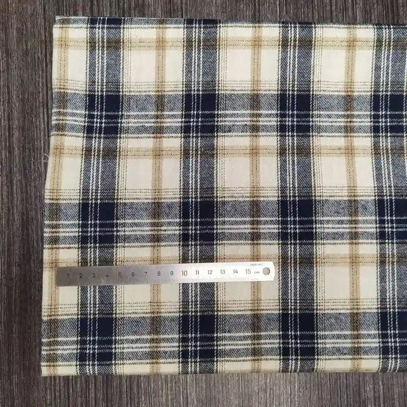 Un morceau de tissu flanelle à carreaux polyester-coton de Longon Craft France, à carreaux beige, marine et blanc, est posé sur du bois avec une règle métallique de 15 cm pour l'échelle. Idéal pour les vêtements d'automne des bébés et vendu au mètre.