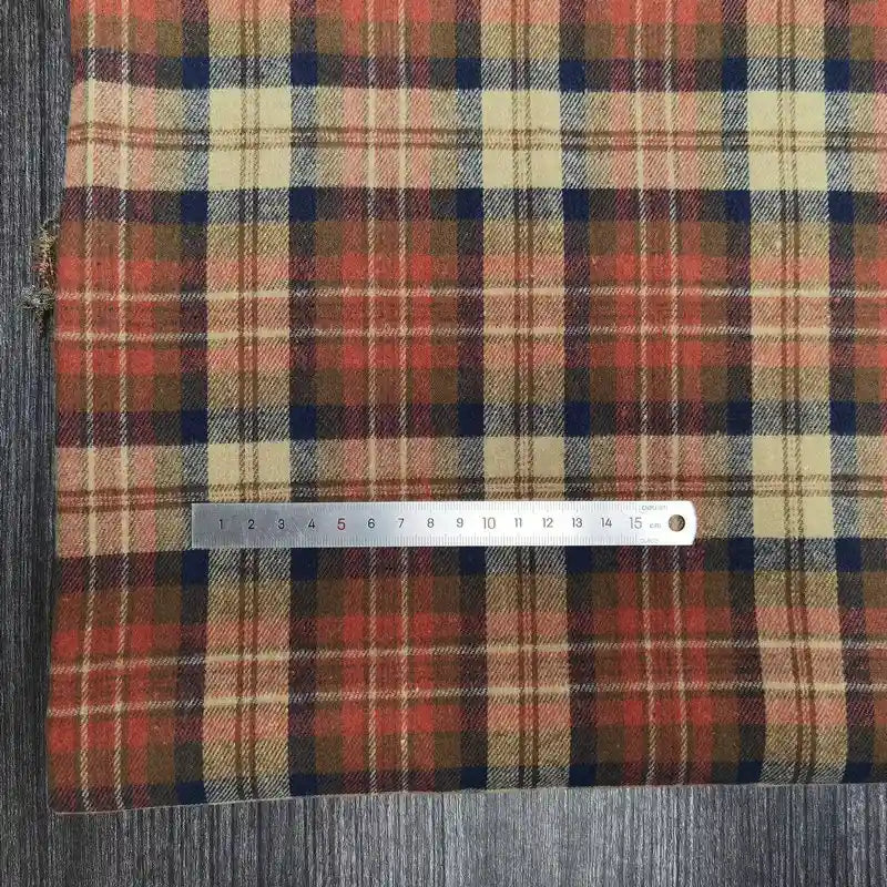 Un tissu rayé rouge, beige et bleu foncé, le tissu flanelle à carreaux polyester-coton de Longon Craft France, est posé sur du bois, avec une règle en métal (0-16 cm) placée sur ce tissu d'automne confortable pour la couture de vêtements de bébé.