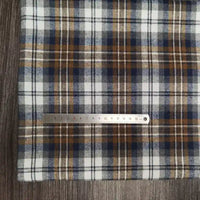 Le tissu Longon Craft France Tissu flanelle à carreaux polyester-coton est présenté sur une surface en bois, avec une règle métallique mesurant environ 20 cm placée horizontalement sur le tissu rayé blanc, bleu, marron et gris.