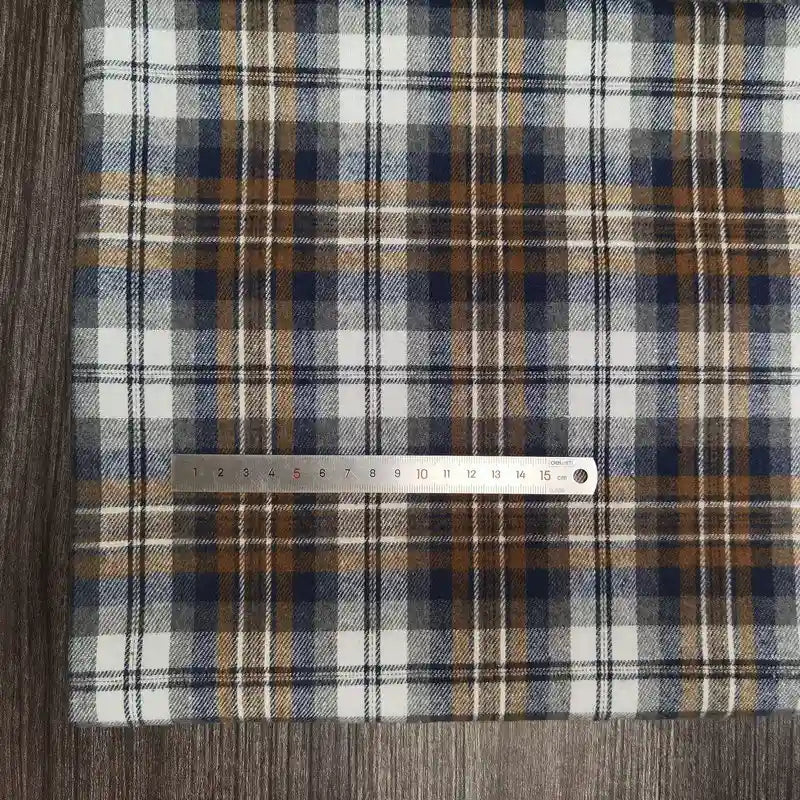 Le tissu Longon Craft France Tissu flanelle à carreaux polyester-coton est présenté sur une surface en bois, avec une règle métallique mesurant environ 20 cm placée horizontalement sur le tissu rayé blanc, bleu, marron et gris.