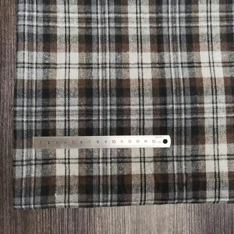 Une règle métallique avec des marques en centimètres et en millimètres est placée à côté du tissu flanelle à carreaux polyester-coton au mètre de Longon Craft France, idéal pour l'automne et la couture de vêtements bébé, présenté sur une surface en bois.