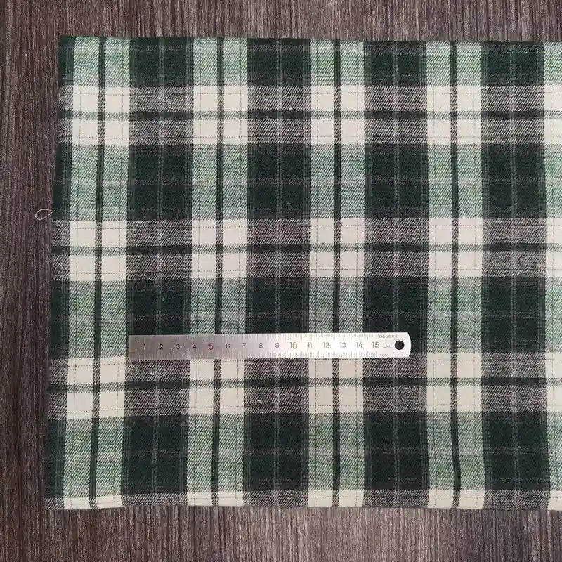 Une règle métallique de 15 cm repose sur le tissu polyester-coton flanelle à carreaux de Longon Craft France, un tissu écossais confortable pour les vêtements d'automne et de bébé, présenté en vert, noir et blanc sur une surface texturée en bois.