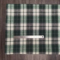 Une règle métallique de 15 cm repose sur le tissu polyester-coton flanelle à carreaux de Longon Craft France, un tissu écossais confortable pour les vêtements d'automne et de bébé, présenté en vert, noir et blanc sur une surface texturée en bois.