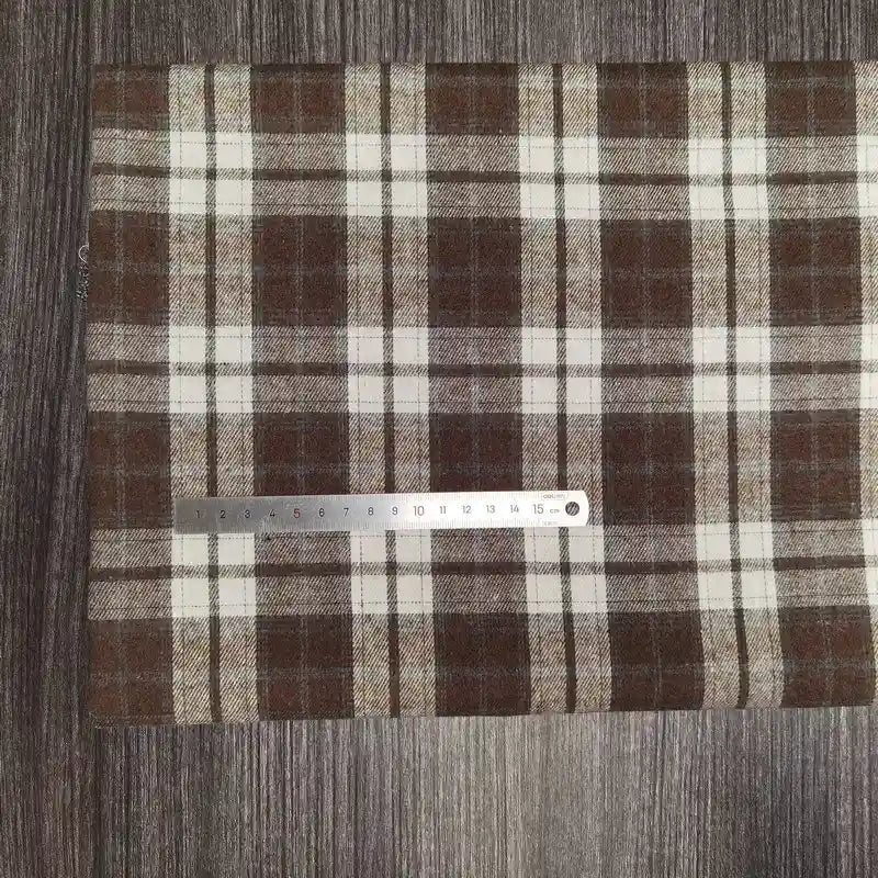 Une règle en métal est posée sur le tissu flanelle à carreaux polyester-coton au mètre de Longon Craft France, un tissu confortable pour les vêtements d'automne et de bébé, présenté sur une surface texturée en bois sombre.