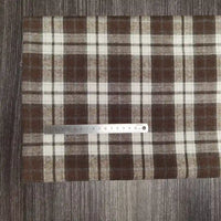 Une règle en métal est posée sur le tissu flanelle à carreaux polyester-coton au mètre de Longon Craft France, un tissu confortable pour les vêtements d'automne et de bébé, présenté sur une surface texturée en bois sombre.
