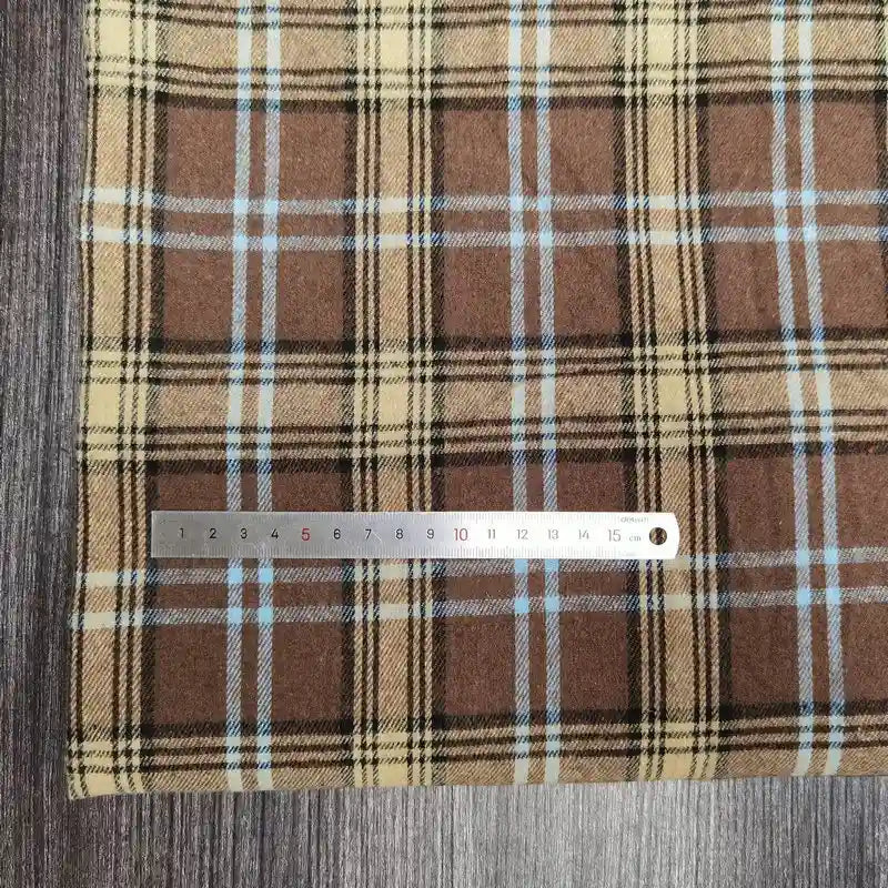 Une règle en métal est posée sur le Tissu flanelle à carreaux polyester-coton de Longon Craft France, une flanelle confortable pour l'automne et les vêtements de bébé, avec des rayures beiges, noires et bleues, placée sur une surface texturée en bois.