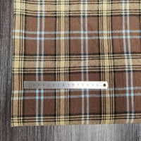 Une règle en métal est posée sur le Tissu flanelle à carreaux polyester-coton de Longon Craft France, une flanelle confortable pour l'automne et les vêtements de bébé, avec des rayures beiges, noires et bleues, placée sur une surface texturée en bois.
