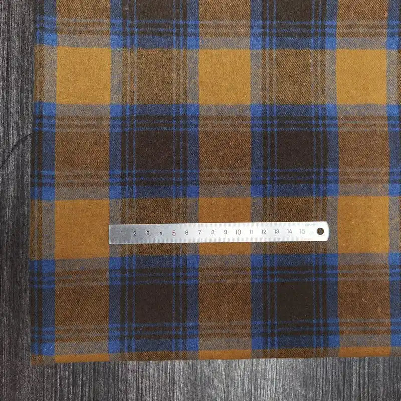 A piece of Longon Craft France tissu flanelle à carreaux polyester-coton, parfait pour vêtements bébé et l'automne, avec motifs jaunes, bruns et bleus, est posé sur du bois. Une règle de 15 cm repose dessus.