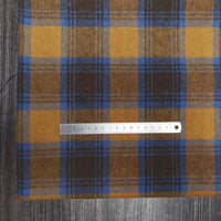 A piece of Longon Craft France tissu flanelle à carreaux polyester-coton, parfait pour vêtements bébé et l'automne, avec motifs jaunes, bruns et bleus, est posé sur du bois. Une règle de 15 cm repose dessus.