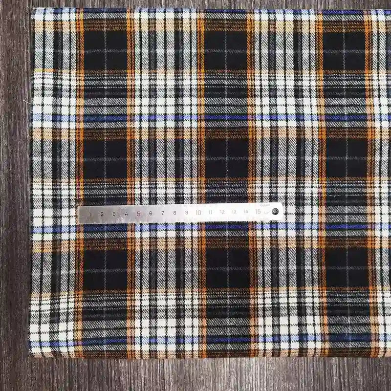 A tissu flanelle à carreaux polyester-coton Longon Craft France, confortable pour l'automne et idéal pour vêtements bébé, avec des rayures noires, blanches, orange et bleues, est posé à plat sur du bois avec une règle argentée dessus pour l'échelle.