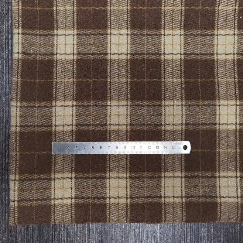 Une règle en métal de 15 cm repose horizontalement sur le Tissu flanelle à carreaux polyester-coton de Longon Craft France, un tissu d'automne confortable pour la couture de vêtements de bébé, dans les coloris marron et beige, posé sur une surface texturée en bois gris.