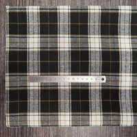 Un morceau rectangulaire de tissu flanelle à carreaux polyester-coton Longon Craft France en noir, blanc et jaune repose sur une surface grise avec une règle argentée en travers - idéal pour la couture de vêtements bébé et les créations d'automne.