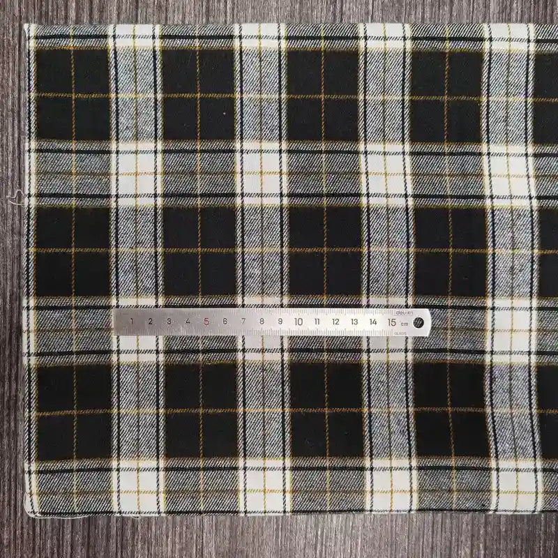 Un morceau rectangulaire de tissu flanelle à carreaux polyester-coton Longon Craft France en noir, blanc et jaune repose sur une surface grise avec une règle argentée en travers - idéal pour la couture de vêtements bébé et les créations d'automne.