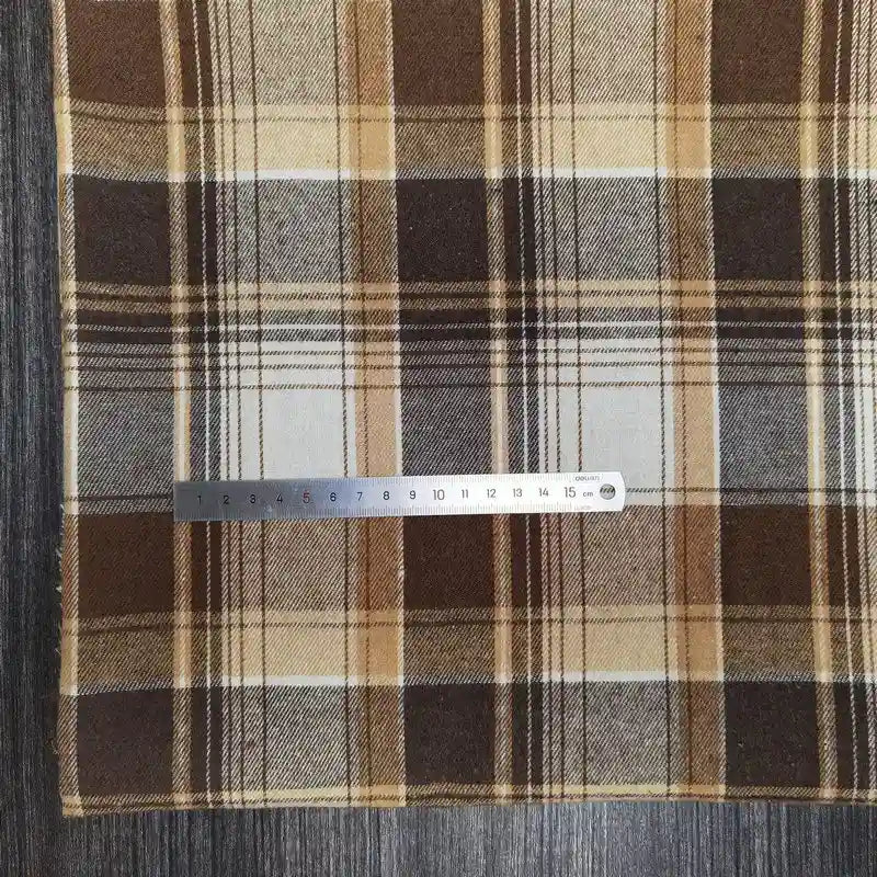 Une règle métallique en centimètres repose sur le tissu flanelle à carreaux polyester-coton au mètre de Longon Craft France, un tissu douillet idéal pour les vêtements d'automne des bébés, sur une surface texturée grise.