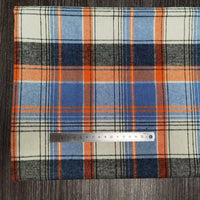 Un Tissu flanelle à carreaux polyester-coton Longon Craft France bleu, orange, blanc et noir est posé sur du bois foncé avec une règle argentée de 15 cm-idéal tissu flanelle confortable pour l'automne et pour vêtements bébé.