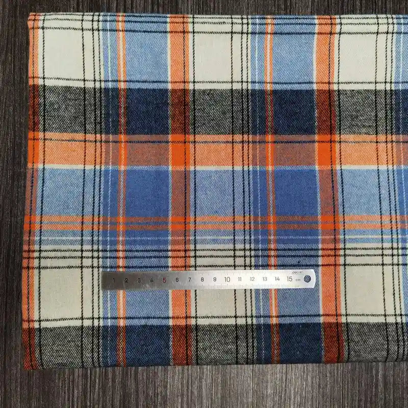 Un Tissu flanelle à carreaux polyester-coton Longon Craft France bleu, orange, blanc et noir est posé sur du bois foncé avec une règle argentée de 15 cm-idéal tissu flanelle confortable pour l'automne et pour vêtements bébé.