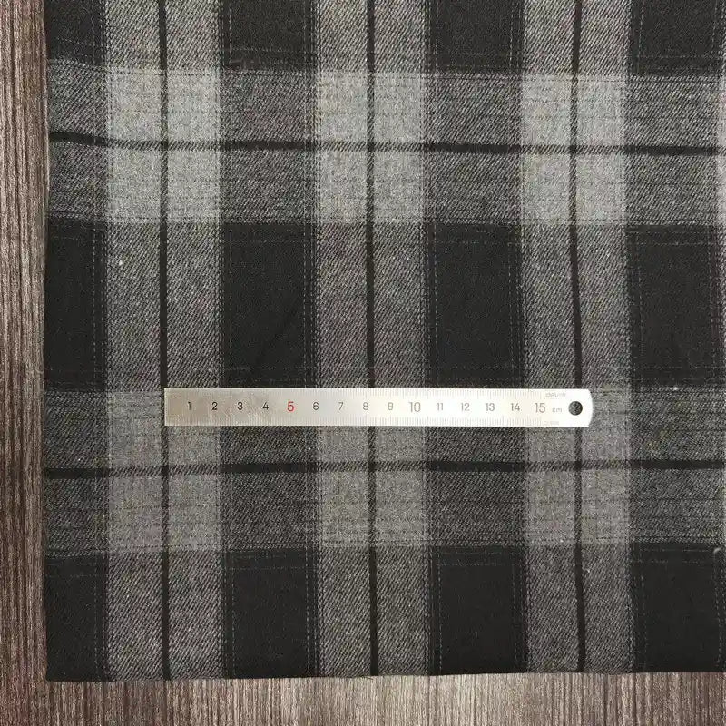 Une règle en métal mesure Tissu flanelle à carreaux polyester-coton au mètre de Longon Craft France en plaid noir-gris de 1 à 15 cm. Cette flanelle douce est parfaite pour l'automne, idéale pour les vêtements de bébé, et présentée sur une surface texturée en bois.