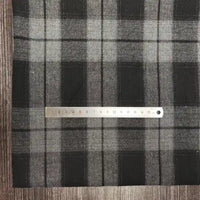 Une règle en métal mesure Tissu flanelle à carreaux polyester-coton au mètre de Longon Craft France en plaid noir-gris de 1 à 15 cm. Cette flanelle douce est parfaite pour l'automne, idéale pour les vêtements de bébé, et présentée sur une surface texturée en bois.