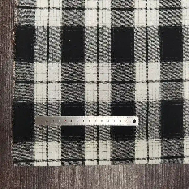Une règle argentée est posée sur le tissu flanelle à carreaux polyester-coton de Longon Craft France, un tissu d'automne douillet vendu au mètre - idéal pour coudre les vêtements des bébés - présenté sur une surface en bois sombre à carreaux noirs et blancs.