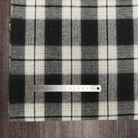 Une règle argentée est posée sur le tissu flanelle à carreaux polyester-coton de Longon Craft France, un tissu d'automne douillet vendu au mètre - idéal pour coudre les vêtements des bébés - présenté sur une surface en bois sombre à carreaux noirs et blancs.