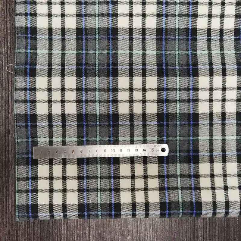 Une règle en métal (18 cm) est posée horizontalement sur le tissu flanelle à carreaux polyester-coton de Longon Craft France, un tissu douillet pour les vêtements d'automne et de bébé, dont les lignes noires, blanches, bleues et vertes sont disposées sur une surface en bois sombre.
