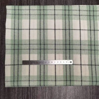 Une règle argentée de 15 cm est posée horizontalement sur le tissu flanelle en polyester-coton vert et beige de Longon Craft France, idéal pour les vêtements d'automne et de bébé, présenté sur une surface en bois sombre.