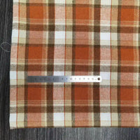 Une règle en métal de Longon Craft France repose sur un tissu flanelle à carreaux polyester-coton au mètre, orange, marron et blanc, idéal pour couture de vêtements bébé et parfait pour l'automne. La règle mesure environ 15 cm.