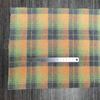 Un morceau de tissu flanelle à carreaux polyester-coton au mètre jaune, vert et bleu de Longon Craft France est posé sur une surface en bois gris, avec une règle métallique de 15 cm placée en travers pour l'échelle.