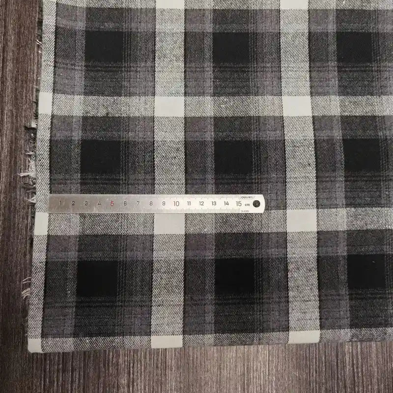 Une règle en métal de 15 cm repose sur le tissu confortable en flanelle de polyester-coton pour vêtements de bébé de Longon Craft France, qui présente un motif à carreaux noirs, blancs et gris avec des bords frangés, posé sur une surface en bois.