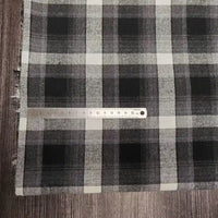 Une règle en métal de 15 cm repose sur le tissu confortable en flanelle de polyester-coton pour vêtements de bébé de Longon Craft France, qui présente un motif à carreaux noirs, blancs et gris avec des bords frangés, posé sur une surface en bois.