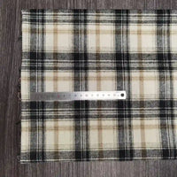 Une règle en métal de 15 cm est posée sur le tissu flanelle à carreaux polyester-coton de Longon Craft France, un tissu d'automne confortable pour les vêtements de bébé, en beige, noir et blanc sur une surface brune texturée.