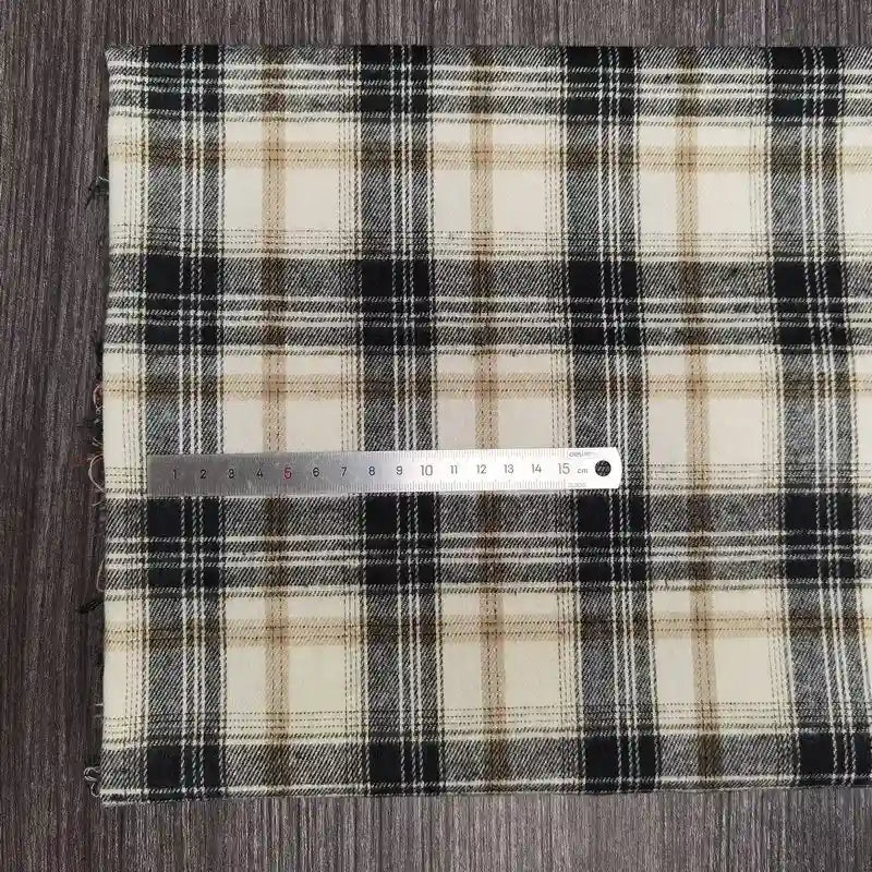 Une règle en métal de 15 cm est posée sur le tissu flanelle à carreaux polyester-coton de Longon Craft France, un tissu d'automne confortable pour les vêtements de bébé, en beige, noir et blanc sur une surface brune texturée.