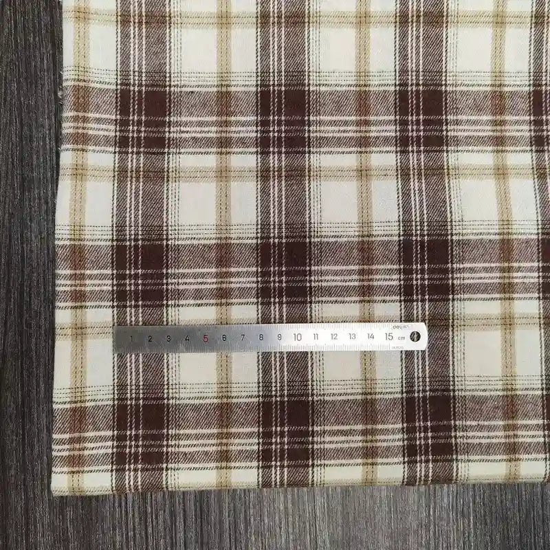 Un tissu flanelle à carreaux polyester-coton de Longon Craft France, avec des rayures brunes, beiges et blanches, est présenté sur une surface en bois. Une règle argentée de 15 cm est posée horizontalement en bas pour l'échelle. Parfait pour vêtements bébé et projets d'automne.