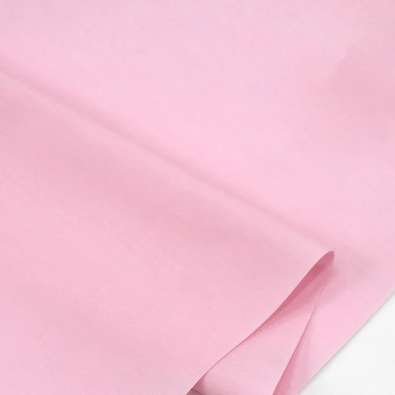 Gros plan sur le Popeline polyester-coton au mètre de Longan Craft France, un tissu popeline à armure toile, doux et respirant, de couleur rose pâle, partiellement plié sur un fond blanc. Convient pour les robes, les chemises et les nappes.
